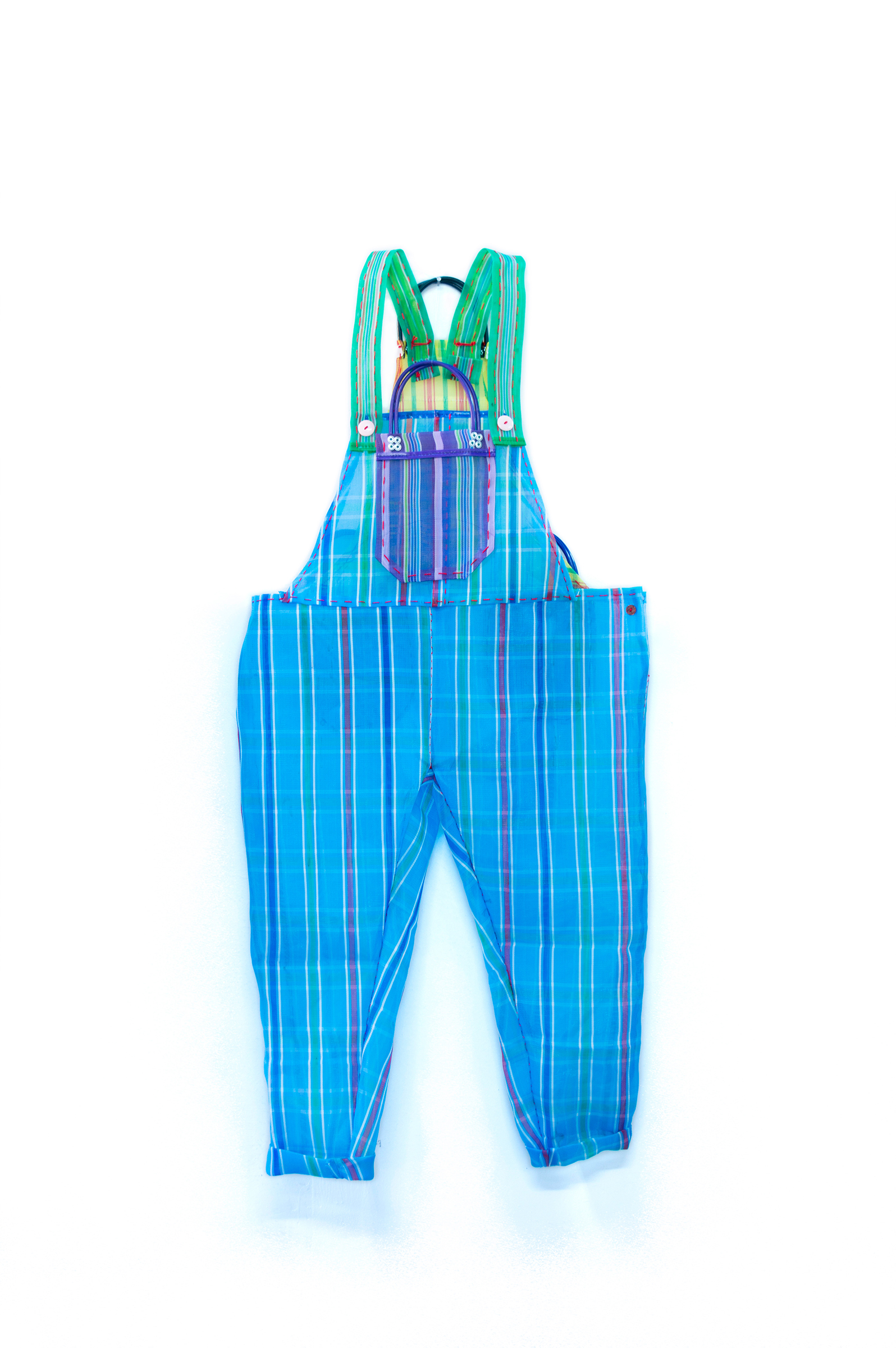 This photograph features a pair of hand-stitched blue overalls with a purple front pocket and green shoulder straps. The overalls are constructed from striped market bags, and the handles of the bags are incorporated into the back of the overalls and the top of the front pocket. The overalls appear against a simple white background.