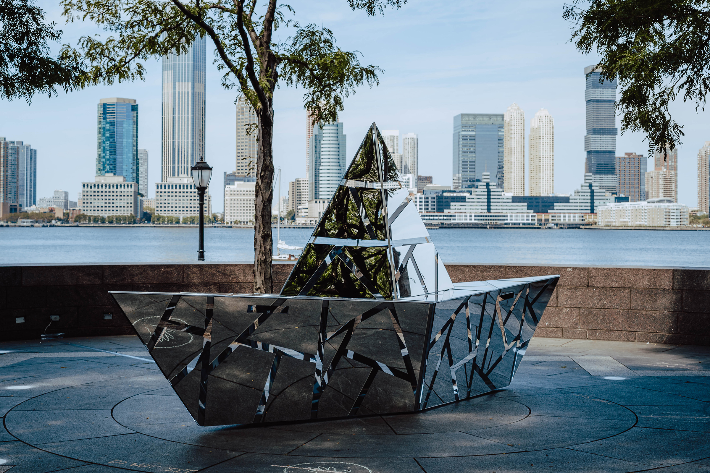 This photograph shows a large metal sculpture resembling an origami boat. The boat is constructed from geometric mirrored panels. There are three-inch spaces between the mirrored panels, so that the geometric shapes appear to float. The sculpture sits on a concrete plaza. Behind it is a low wall, a tree, and a river, with an urban skyline on the far side of the river. The sky is light blue with wispy clouds.