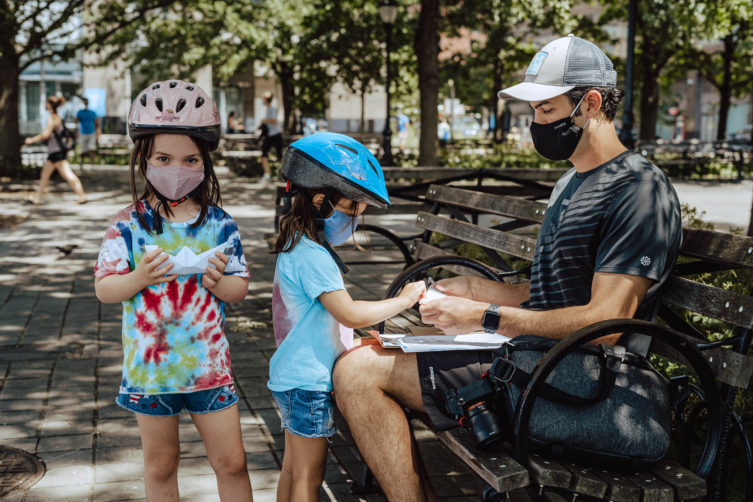 This photograph shows a young family in an urban park. The father is seated on a park bench on the right side of the picture. He wears shorts, a T-shirt, a baseball cap, and a facemask. On his lap he holds a stack of white papers. His backpack and camera rest on the bench beside him. Standing in front of him are two young girls. One of the girls faces the man and reaches to take a piece of paper. The second girl, on the left side of the photo, holds a completed origami boat and looks directly at the camera. Both of the girls wear denim shorts, tie-dye T-shirts, bike helmets, and facemasks.