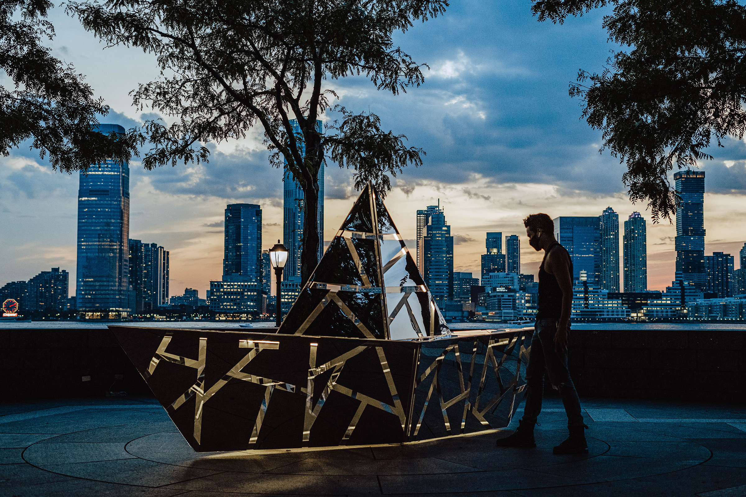 This photograph shows the same sculpture and scene as the previous photo, but this image was taken at dusk. The boat sculpture is illuminated from within with a warm, yellow light. Lights are also on in the buildings in the city skyline on the far side of the river. A man stands to the right of the sculpture, looking down at it. He appears as a dark silhouette against the early evening sky.