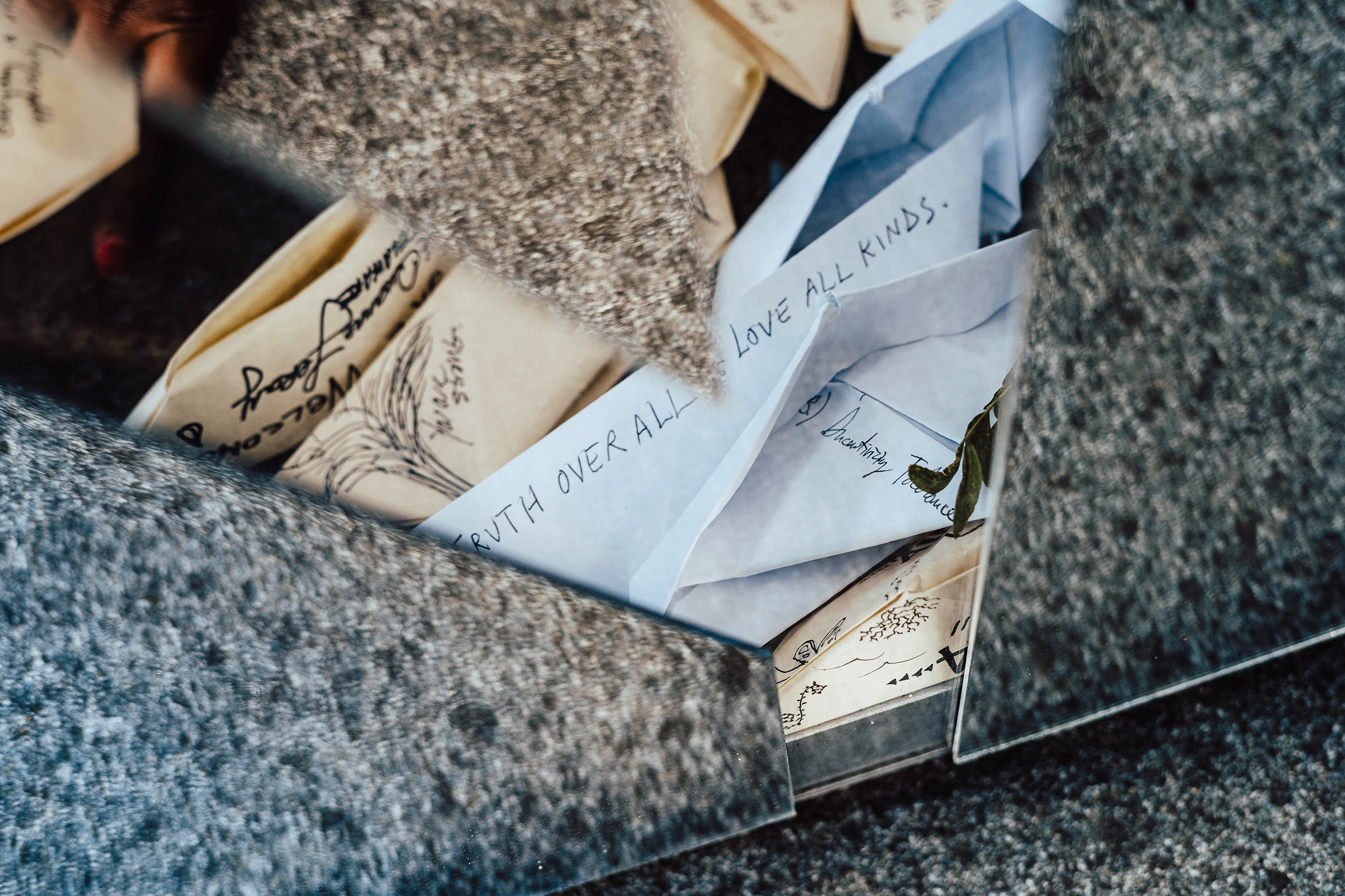 This photograph is a close-up of a section of the large boat sculpture. The image shows a gap between three of the geometric mirrored panels that form the outer structure of the boat. In the gap between the panels, there are several small paper boats inscribed with written notes and drawings. One of the notes in the foreground reads, 'TRUTH OVER ALL. LOVE ALL KINDS.'