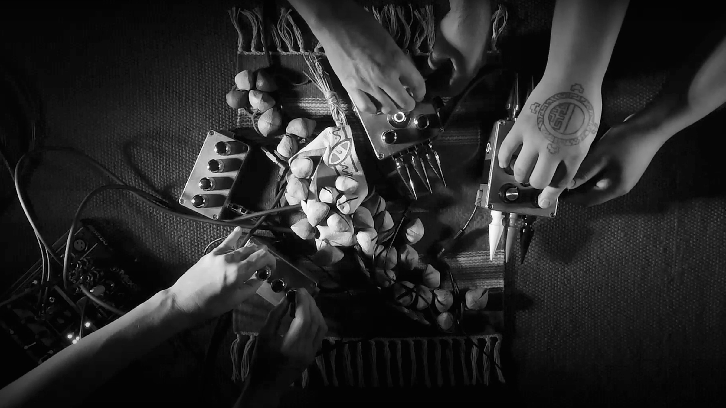 This black-and-white photographic image is a still from a video. It is a top-down view of three pairs of hands playing four electronic instruments. The instruments are boxes with dials and spikes, and they are positioned on top of a small, woven rug with ayoyote rattles in the center.
