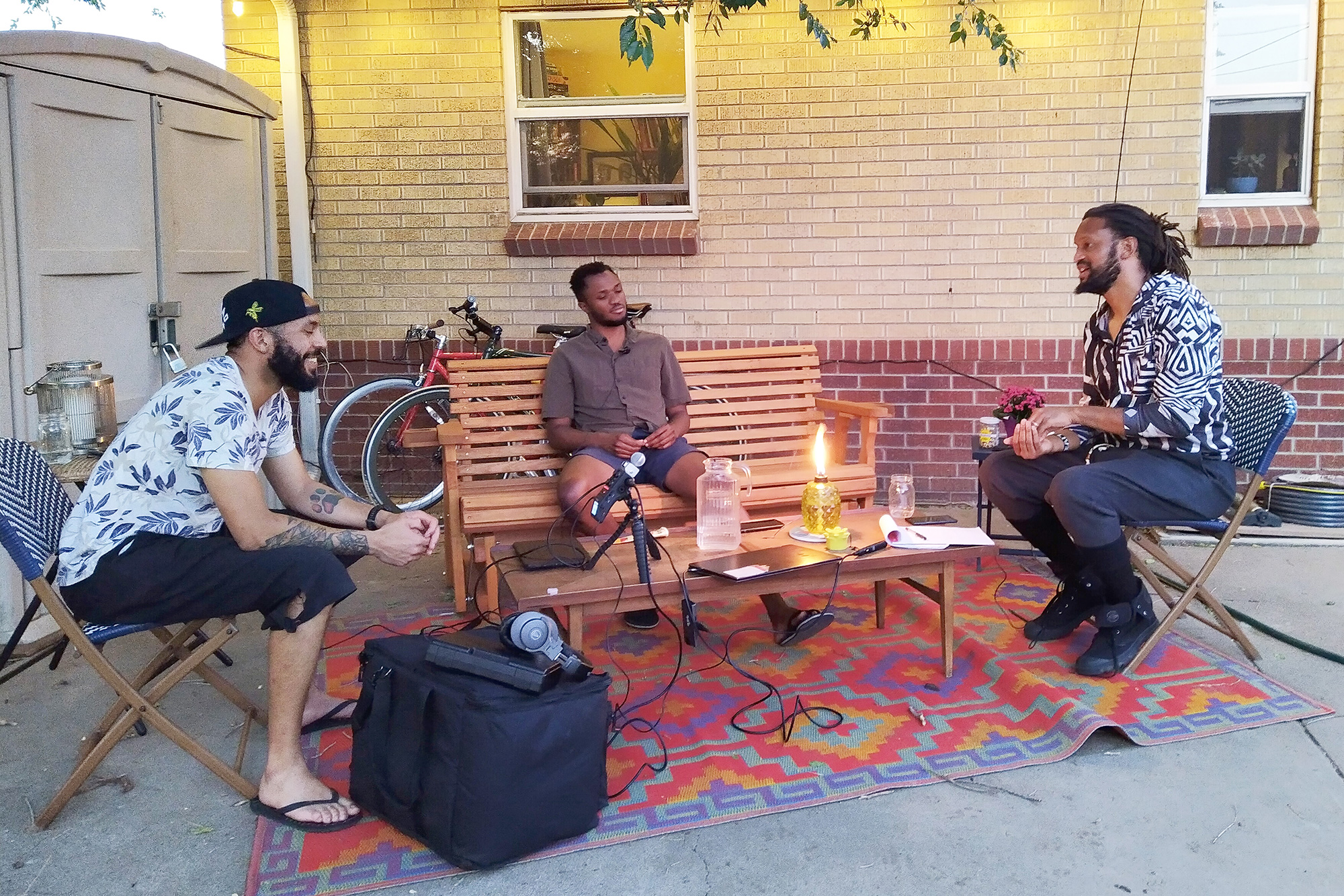 Photo of three young men, seated on outdoor furniture atop a multi-colored rug with recording equipment in evidence. To the left is Cory Minkah Montalvo, a Black, Latino man in a black baseball cap and flip flops; in the middle is Wisdom Amouzou, a man of African descent wearing shorts and a brown shirt, and to the right is Stephen Brackett, an African-American man wearing a black-and-white patterned shirt and shorts.