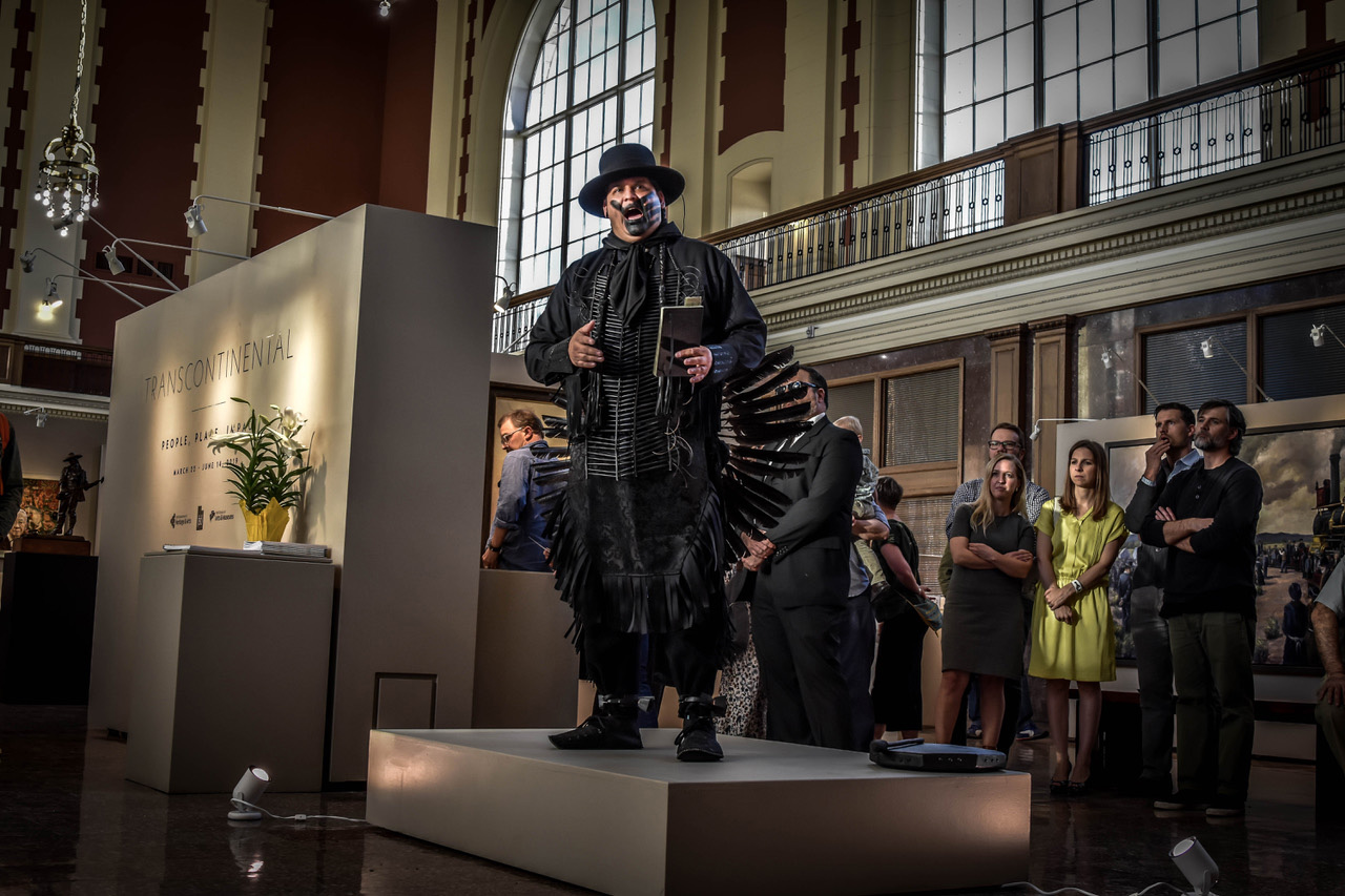 This photograph depicts the artist, Gregg Deal, standing on a low pedestal in a gallery space with high ceilings and arched windows. Viewers stand behind and to the right, watching him. Deal is dressed entirely in black. He wears Indigenous powwow garments, including a hat with a wide brim. His open mouth is obscured by black face paint in the shape of a large handprint. Deal is speaking, and he is gesticulating with his raised hands. His right hand holds a book.