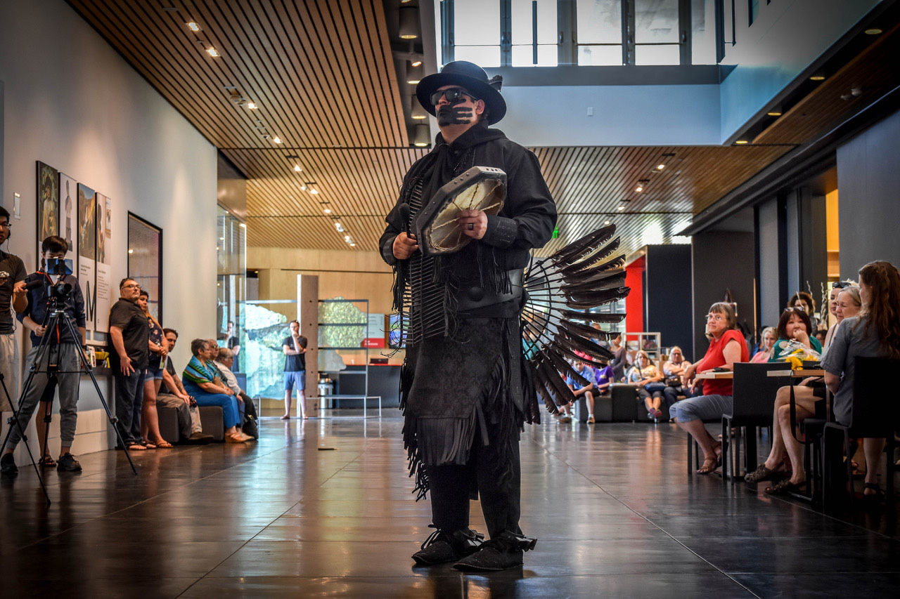 This photograph depicts the artist, Gregg Deal, standing rigidly in the center of a museum lobby. Viewers stand or sit along the walls to the left and right of him. Deal is dressed entirely in black. He wears Indigenous powwow garments, including a hat with a wide brim. His eyes are hidden behind dark glasses. His mouth is obscured by black face paint in the shape of a large handprint. He holds a drum in his left hand and a drum stick in his right hand.
