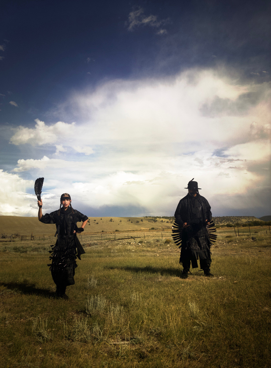 This photograph captures two figures standing in an open field. The blue sky is filled with puffy white clouds, and a fence line runs through the background of the image. Both figures are clothed in Indigenous powwow garments, but all the clothing is black. The figure on the right is artist Gregg Deal. He wears a black hat and stands with his feet apart. His face is obscured by a large black handprint, covering his mouth. The figure on the right is Deal’s teen daughter, Sage. Her hair is braided, and she wears a headband. In her raised right hand, she holds a fan of black feathers. Her left hand rests on her left hip. She looks at the camera. Both figures cast dramatic shadows on the ground.