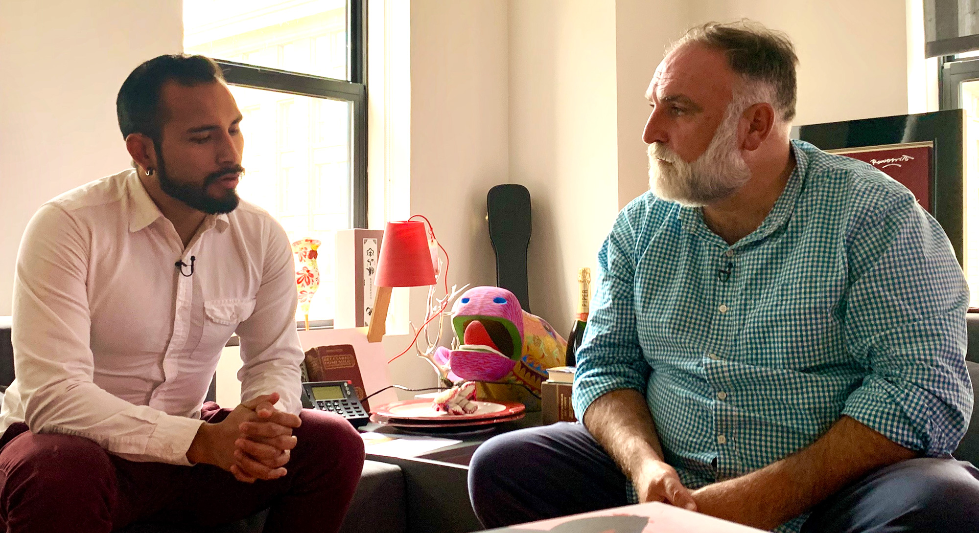 Two men are seated and engaged in conversation. To the left is Alejandro Fuentes-Mena, a young man of Chilean descent with a tightly-cropped beard, wearing a white shirt; to the right is chef José Andrés, a middle-aged man with a grey beard wearing a checked green-and-white shirt.