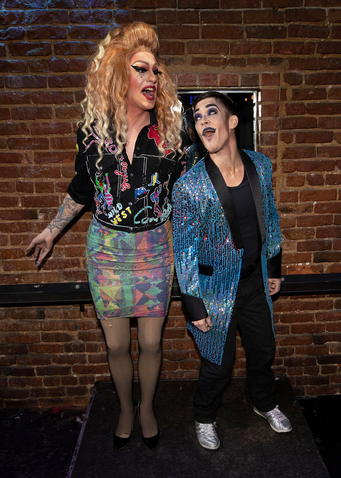 Two drag performers standing together against the backdrop of a brick wall in a performance venue. On the left is Kai Lee Mykels, who has long, curly blonde hair and wears a “Wild West” black denim jacket and a colorful mini-skirt. On the right is Trey Suits, wearing a long, blue, sequined jacket, black pants, and silver sneakers. Both performers have their mouths open, as if they are singing or speaking.