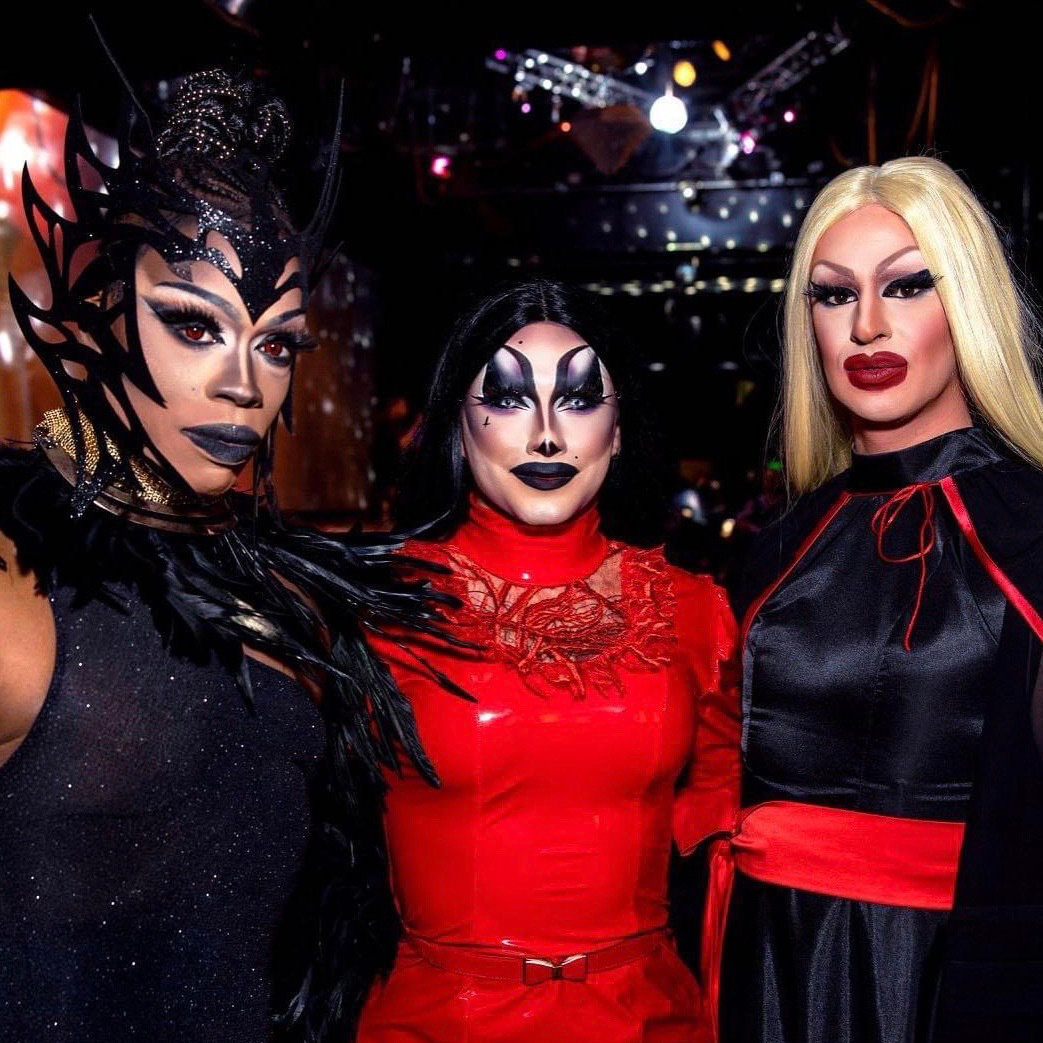 Three drag performers standing together against the backdrop of a performance venue. To the left is Khrys’ta Aal, a Black performer wearing a sparkling, black dress, feathers about the neck, and an elaborate black headdress; in the middle is Veronica Taylor Mykels, who has long, dark hair and wears a bright red dress and cat-eye makeup; and to the right is Kai Lee Mykels, who has long blonde hair and wears a black and red dress and deep red lipstick.