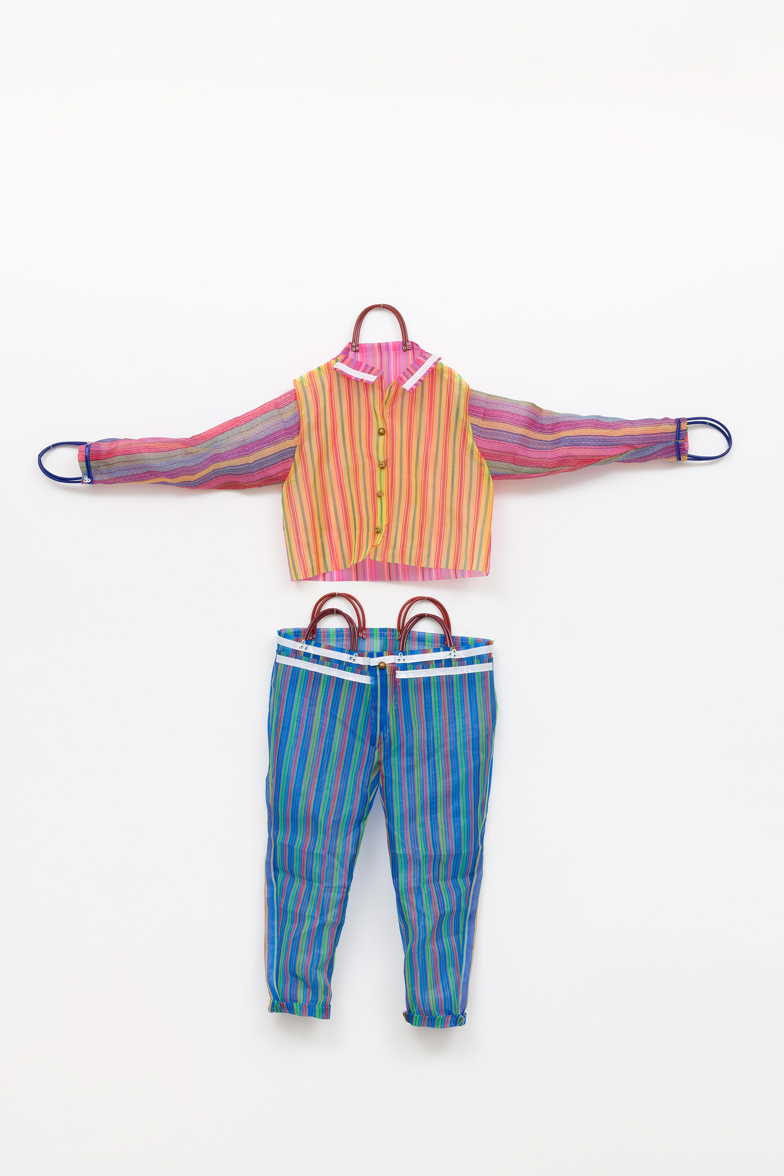 This photograph features a colorful two-piece outfit, including a pink and yellow button-up top and blue pants, both made from striped market bags. The handles of the bags are incorporated into the neckline and sleeves of the shirt and the waistband of the pants. The outfit appears against a simple white background.