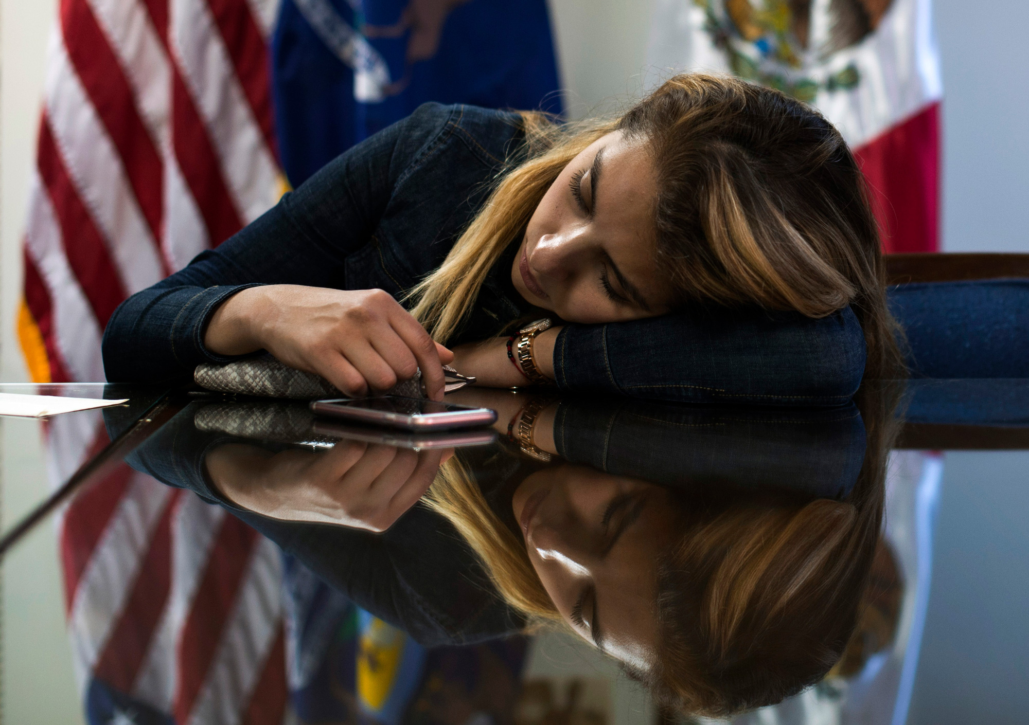 This photograph shows a young woman (Pamela) with her head on her arm resting on a table. She wears a dark denim shirt and looks down at her phone on the table. Behind her, three flags hang from flagpoles. The surface of the table is highly glossy, reflecting Pamela’s image and the flags.