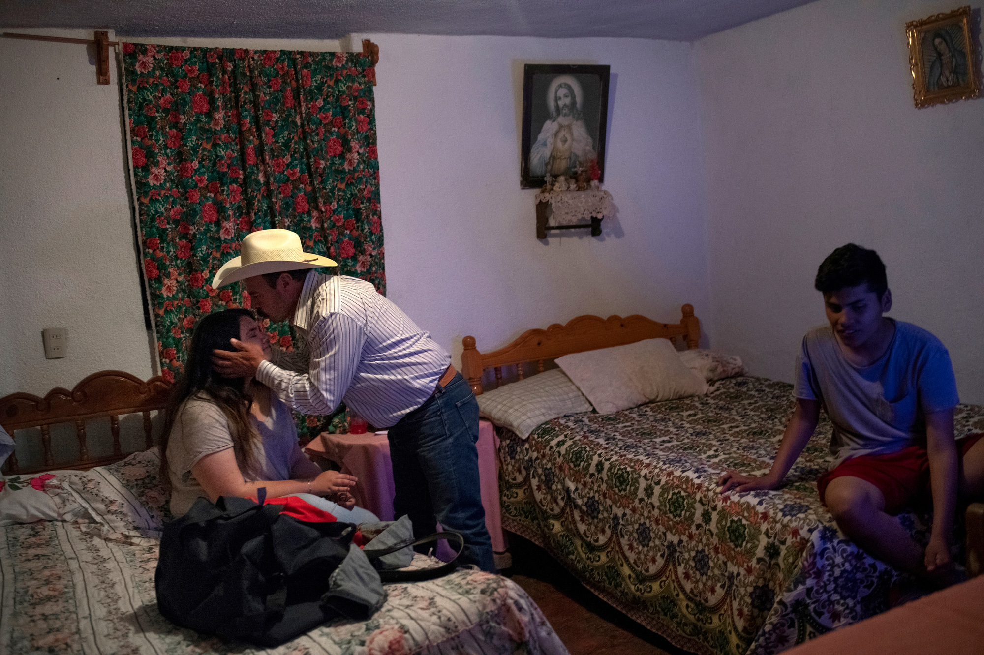 On the left side of this photograph a middle-aged man (Luis) leans down to kiss a teenage girl (Lulys) on her forehead. Lulys is wearing a gray t-shirt, and she is sitting on a twin bed with a floral bedspread. Luis wears a straw cowboy hat, a blue-and-white striped, button-down shirt, and jeans. On the right side of the photograph, a teenage boy (Bryan) sits on another twin bed with a floral bedspread. Bryan wears shorts and a T-shirt. A painting of Jesus hangs above a mounted altar on the wall above Bryan’s bed.