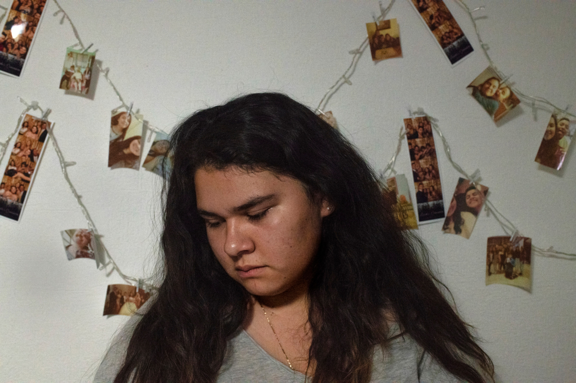 This photograph shows a teenage girl (Lulys) with long, dark hair and downcast eyes. She stands in front of a white wall with printed photographs clipped to strings. The pictures show other teenagers smiling.