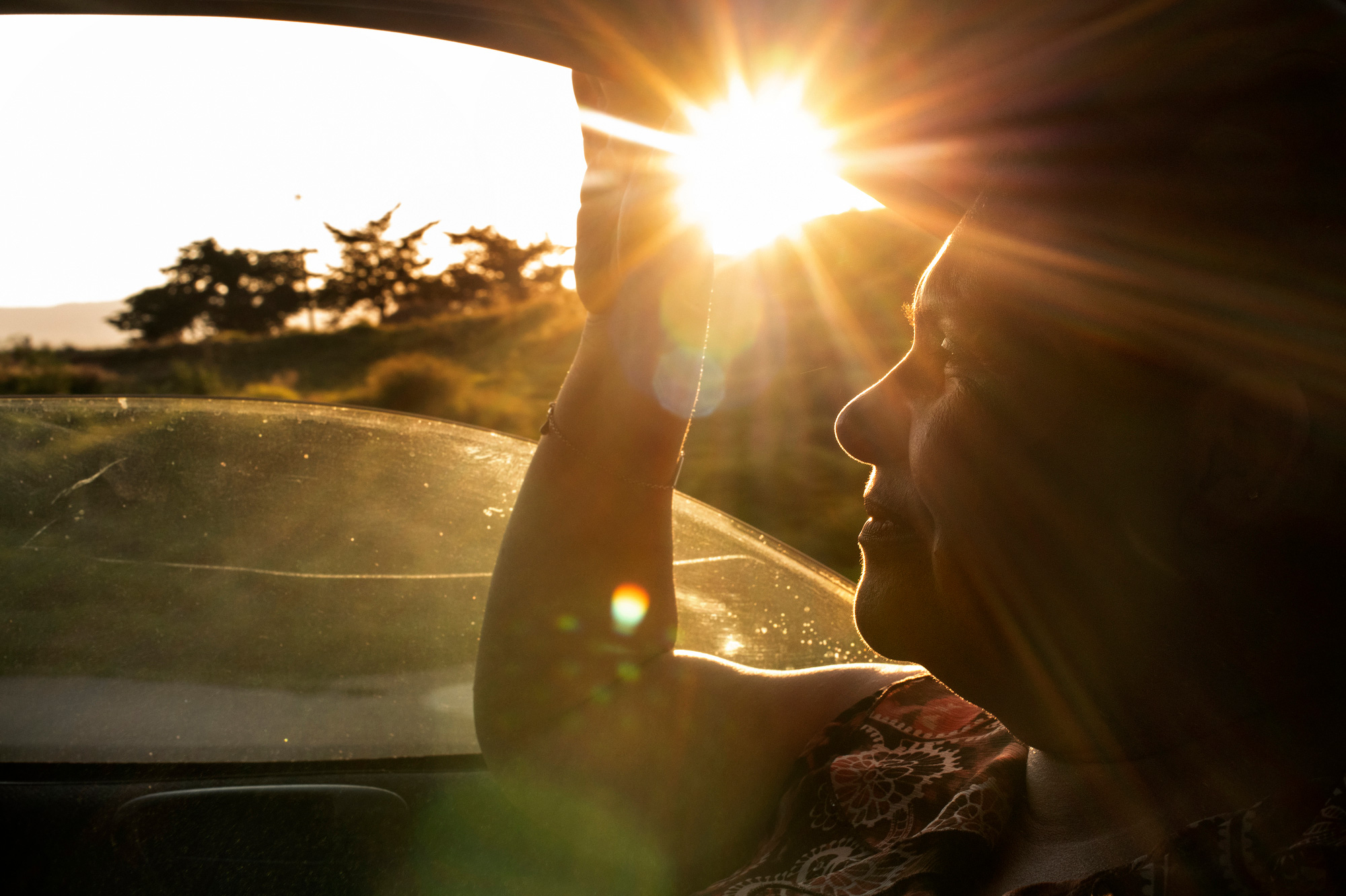 A middle-aged woman (Lourdes) sits in the front passenger seat of a car with the window rolled down. She looks straight ahead to the left side of the image. A golden sunbeam shines in, so her profile is backlit. The car window is rolled down halfway, and Lourdes holds on to the upper window frame with her right hand.