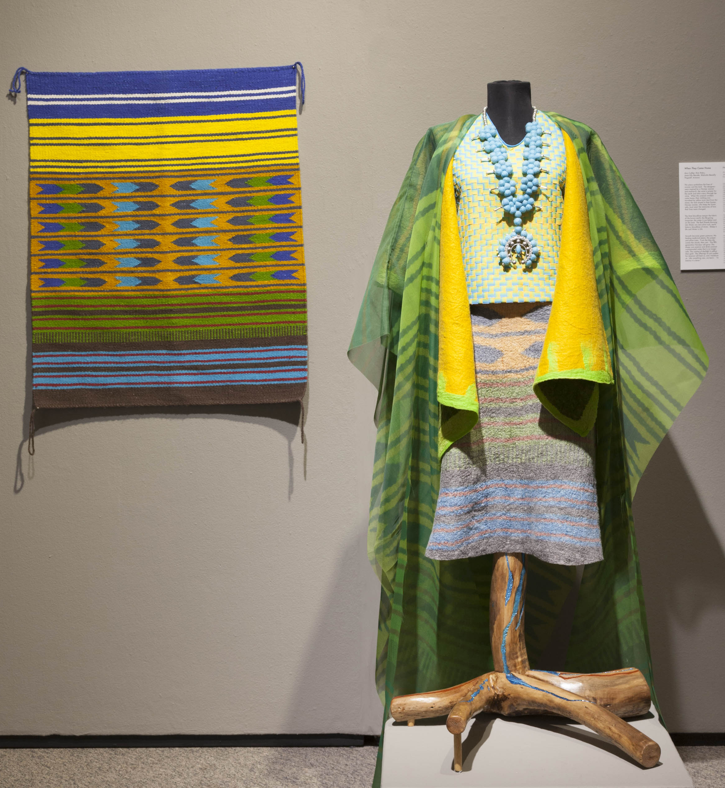 This photograph shows the artists’ works installed in a museum gallery. On the left, a wool rug with stripes of yellow, blue, green, orange, and brown hangs on a wall. To the right is a headless mannequin, dressed in clothing inspired by traditional Navajo designs: a skirt, blouse, cape, shroud, and necklace, all in colors similar to those of the rug.