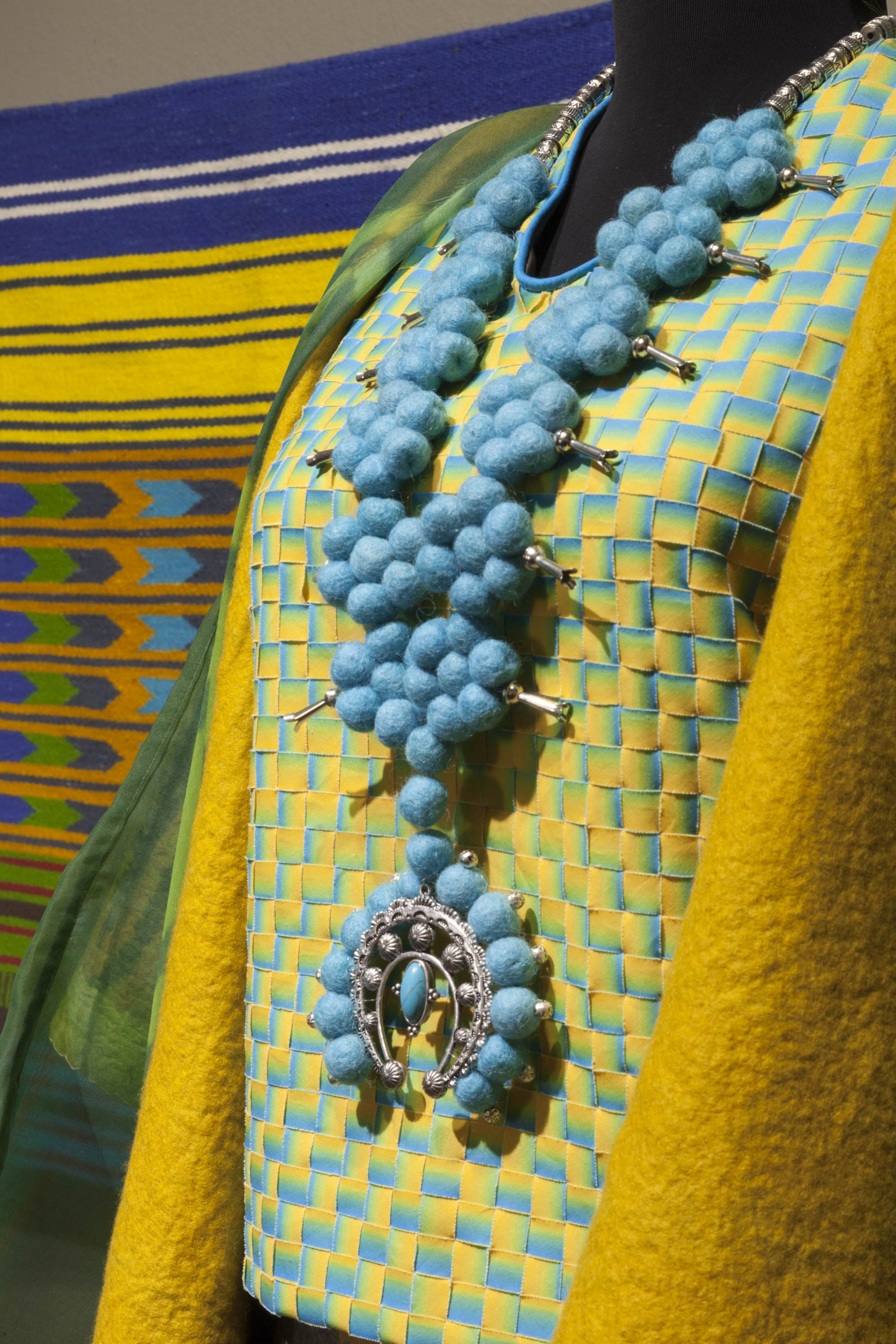 This photograph features a close-up of a necklace and blouse displayed on a mannequin. The necklace, inspired by traditional Navajo designs, is made of felted blue wool balls and silver. The woven blouse is yellow and blue. A wool rug is hanging on the wall behind the mannequin.