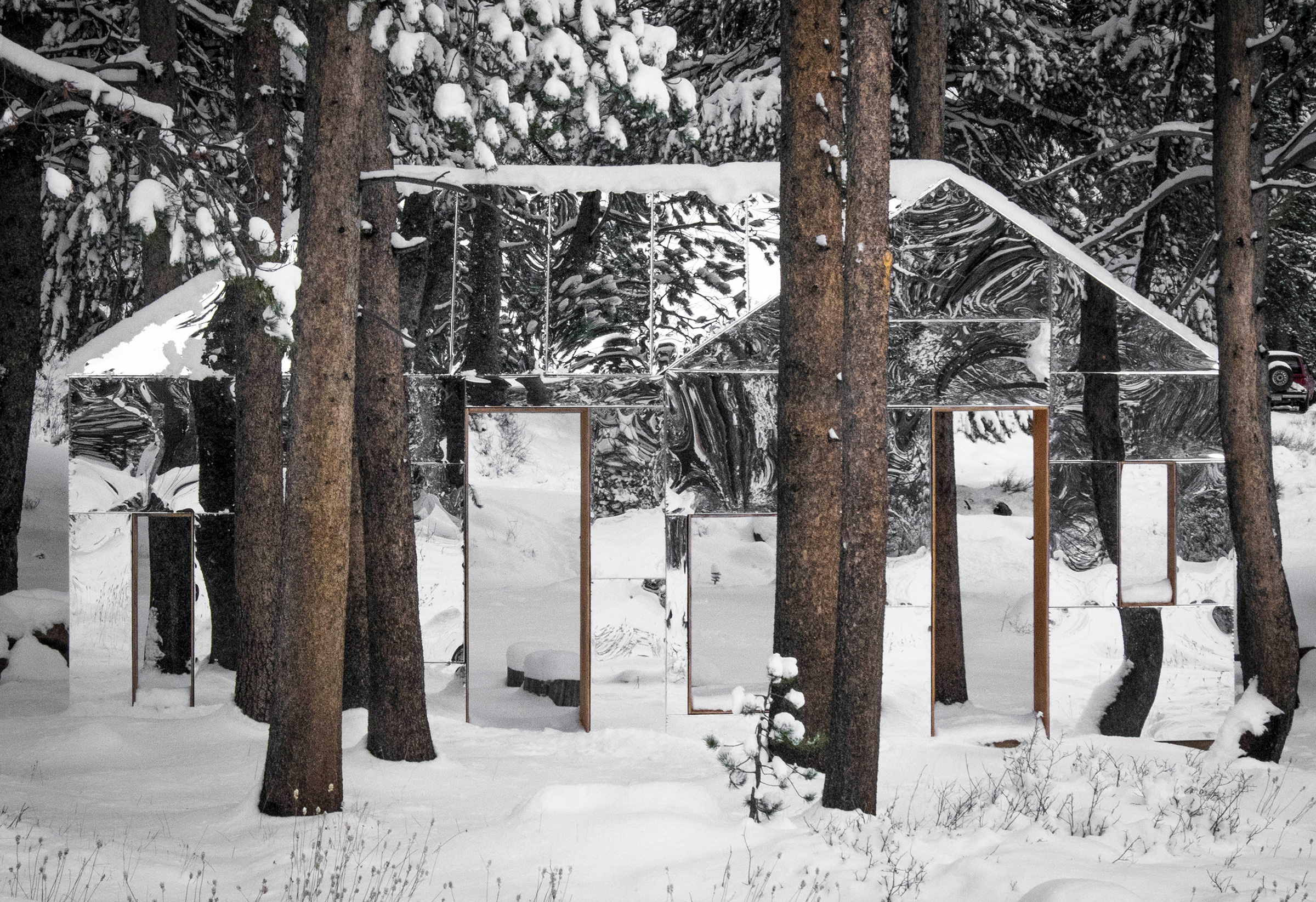 This photograph features a large, mirrored sculpture, set in a wooded, snow-covered area. The sculpture is shaped like a simple house or cabin structure, and the mirrored surface reflects the evergreen trees and snow around it, so the sculpture seems to disappear into the landscape.