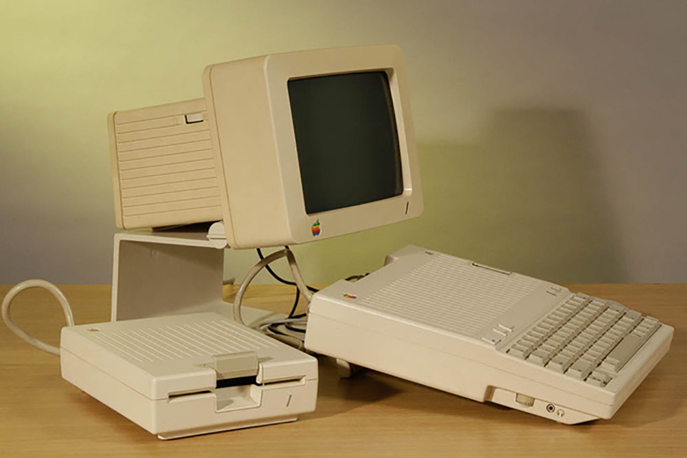 This photograph depicts an Apple IIc computer positioned on top of a wood laminate desk. The computer components (keyboard, display, and disk drive) are all light beige, and a logo with a rainbow-colored apple is prominent below the computer screen.